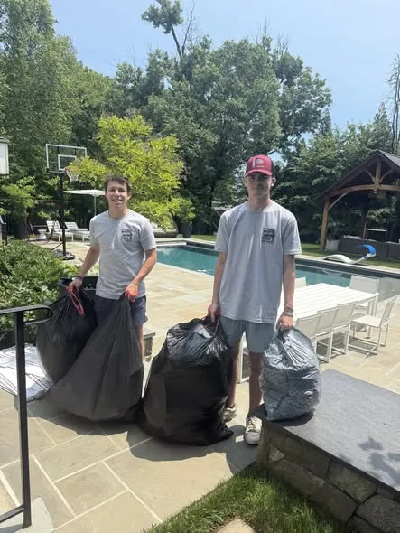 Two crew members carrying bagged junk out of a backyard pool patio cleanup